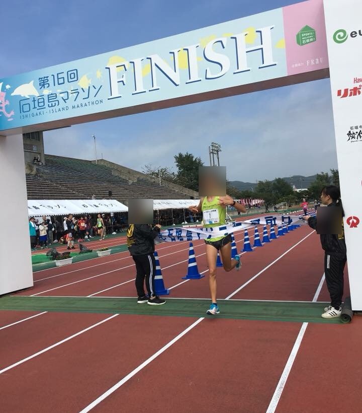 紅色跑道上的終點拱門,門上寫著「第16回 石垣島マラソン FINISH」,一位跑者衝過終點布條,兩側各站一位工作人員,後方是陸上競技場的看台與白色帳篷