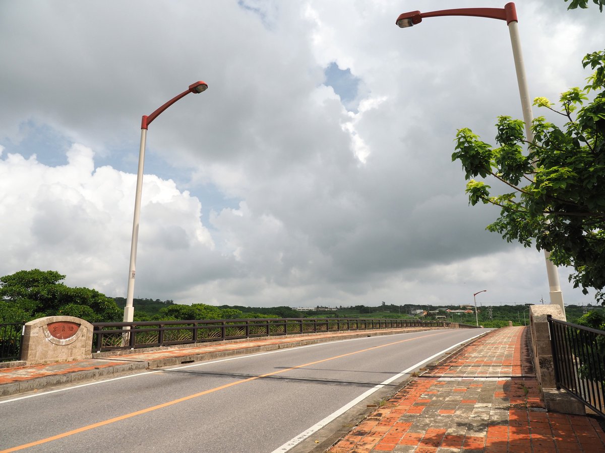國道 390 號的宮良橋:兩線道的柏油路往前延伸,右側是紅磚人行道,橋頭石柱上有紅底的橋名牌,兩旁立著紅色路燈,天上是大片白雲