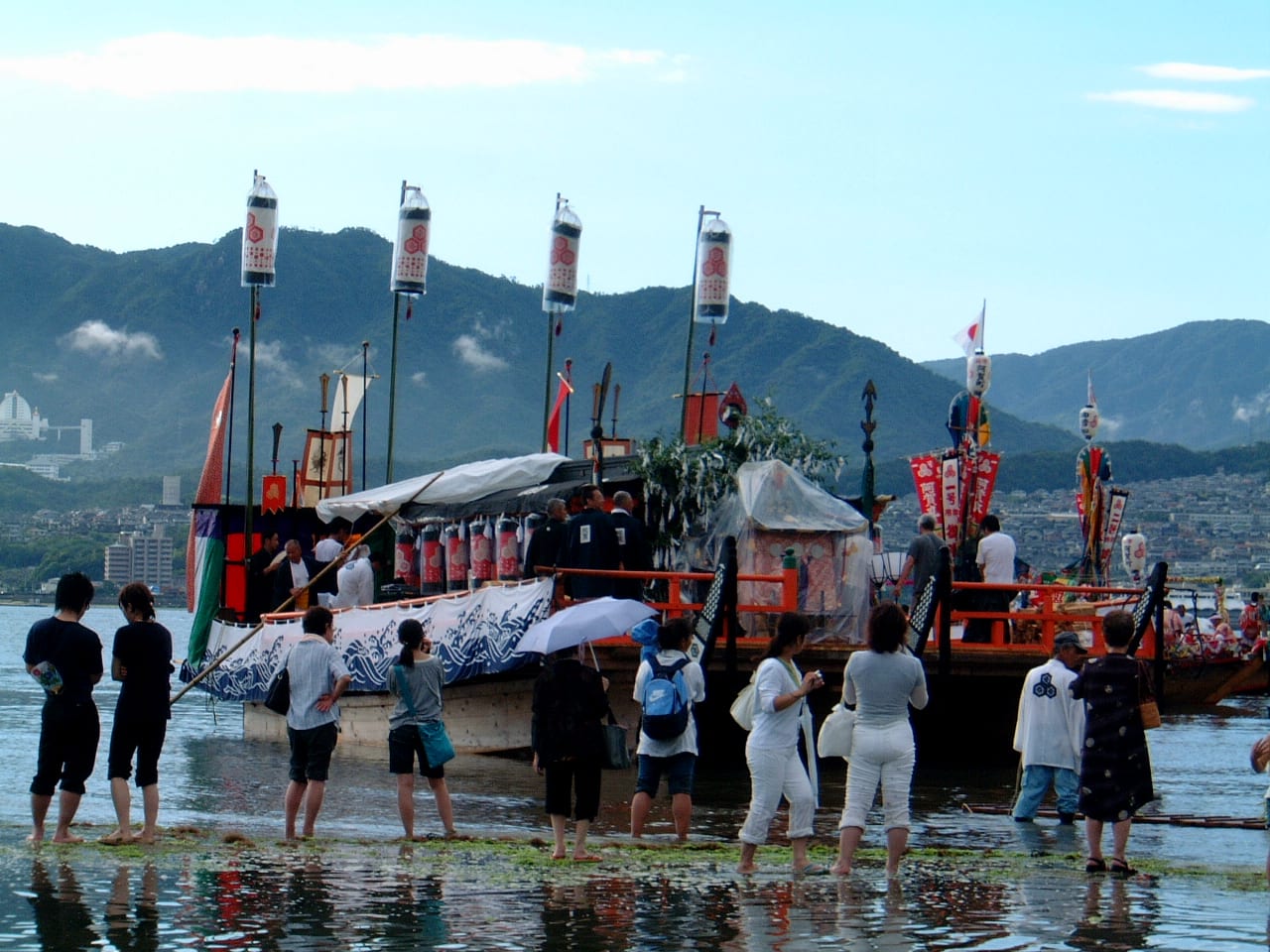 The decorated festival barge of Kangensai moored in the shallows off Miyajima, hung with lanterns and banners, with spectators standing barefoot in the water