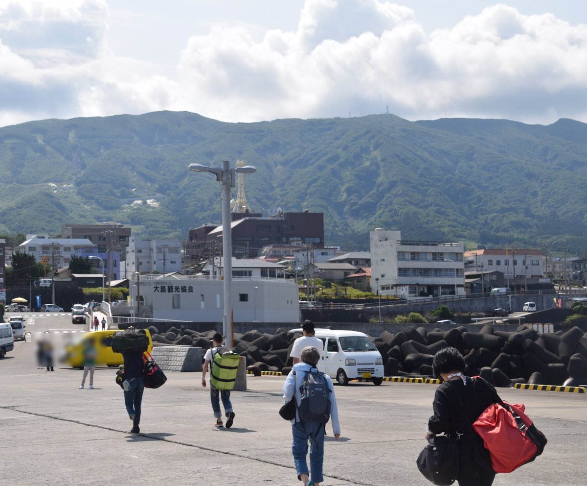 元町港碼頭：剛下船的旅客背著行李往岸上走，前方是白色建築「大島観光協会」與消波塊，後方是元町的樓房，再後面是長滿綠色植被的山坡