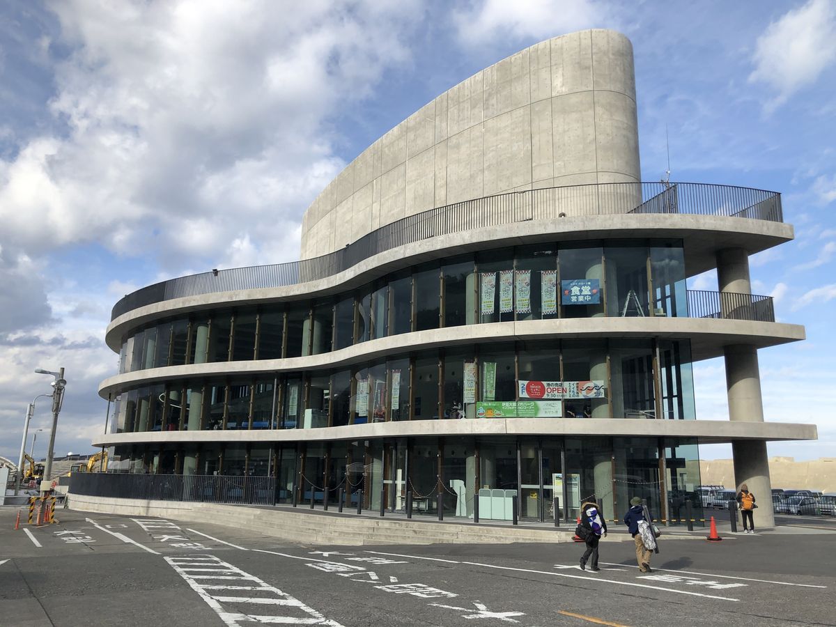 岡田港的船客待合所：清水混凝土的三層弧形建築，每層是一圈玻璃窗與外廊，三樓窗內掛著「食堂 営業中」的看板，前方停車場有兩三位旅客走過