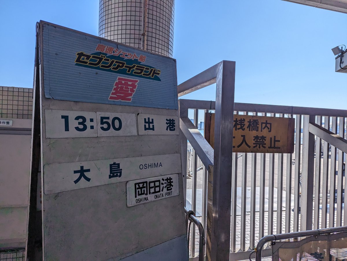 竹芝棧橋乘船口旁的出港告示板：上方寫「高速ジェット船 セブンアイランド 愛」，中間是「13:50 出港」，下面是「大島 OSHIMA」，再下面另外掛著一塊「岡田港 OSHIMA OKATA PORT」的小牌，右側欄杆上掛著「棧橋内 立入禁止」