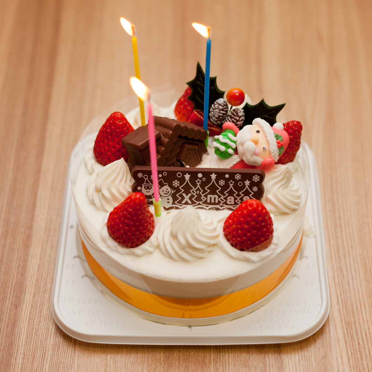 A Japanese Christmas cake: a round white cream sponge topped with whole strawberries, a chocolate X'mas plaque, a sugar Santa figure and three lit candles