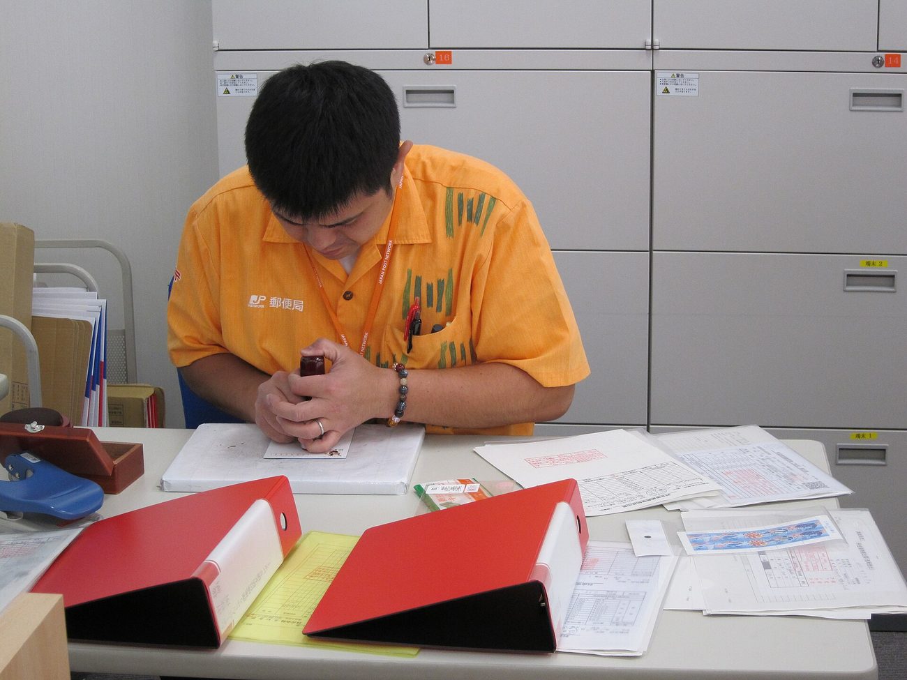 Behind the counter of a Japanese post office, a male clerk in an orange uniform bends over a document to apply a hanko stamp; the JP logo and the characters for post office are embroidered on his chest, two red ring binders, several red-ruled forms and an ink pad lie on the desk, and grey filing cabinets fill the wall behind him