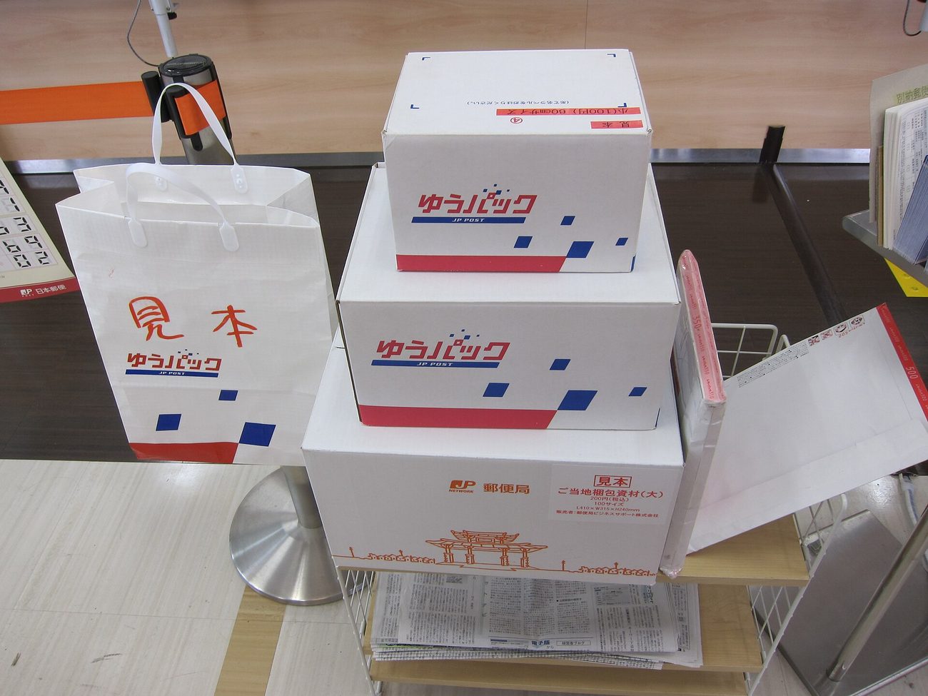 A sample display beside a post office counter holding three Japan Post packing items: a white paper carrier bag stamped in red with the word sample and the Yu-Pack JP POST logo, two white Yu-Pack cartons with red and blue geometric blocks, and a larger box underneath printed with the post office logo, an orange line drawing of a gate, and the wording local packing material, large, 200 yen including tax, with dimensions L410 x W315 x H280 mm