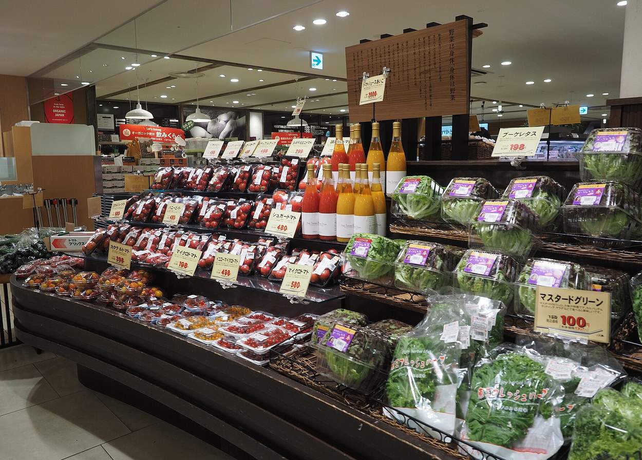 The fresh produce floor of a Japanese supermarket, with tomatoes, salad leaves and bottled juices laid out under handwritten price cards