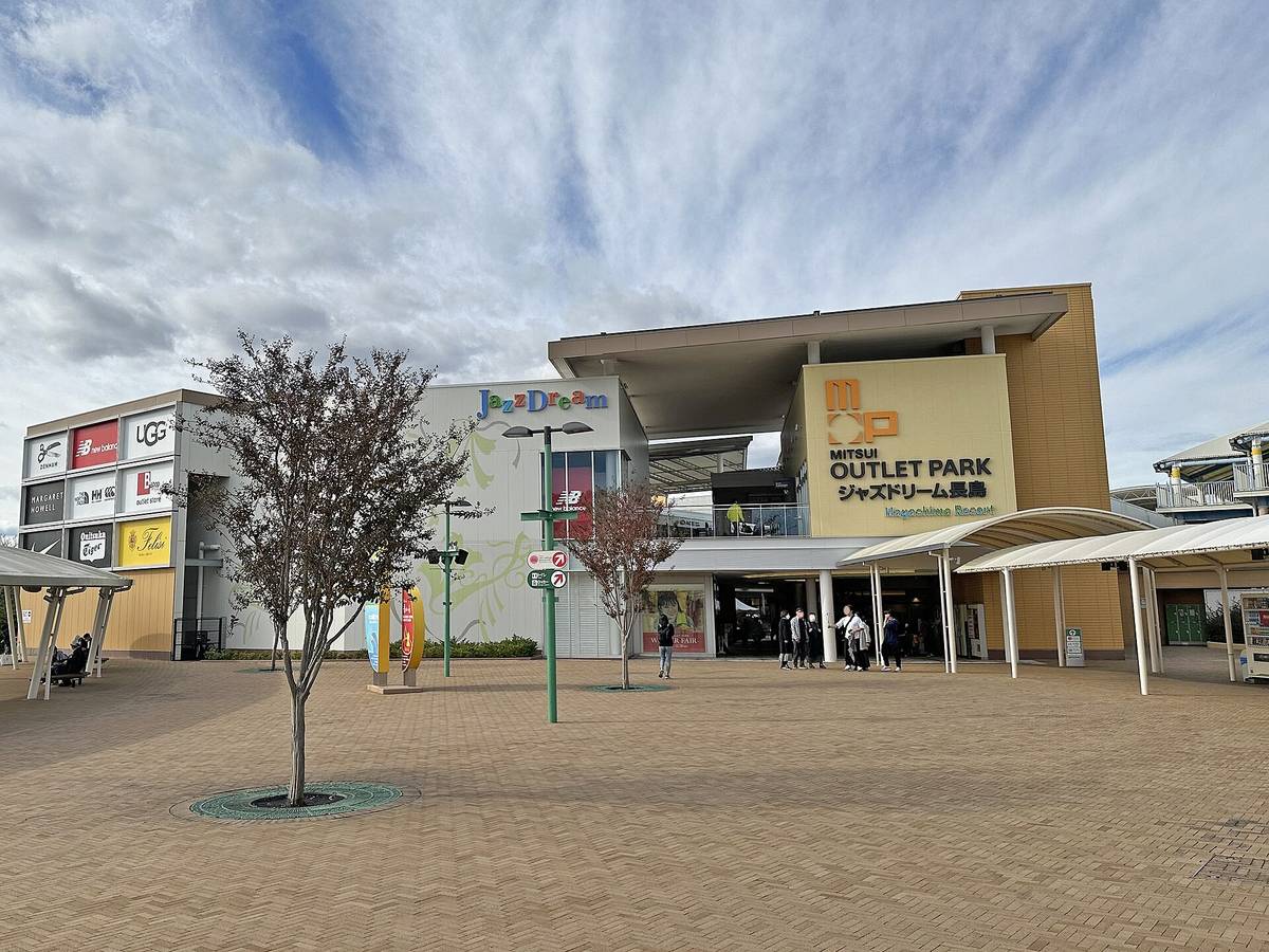 Entrance plaza of Mitsui Outlet Park Jazz Dream Nagashima: a cream facade with the Mitsui Outlet Park lettering and a multicoloured JazzDream logo, brand signs such as UGG and New Balance on the left wall, a brick plaza and white canopies in front