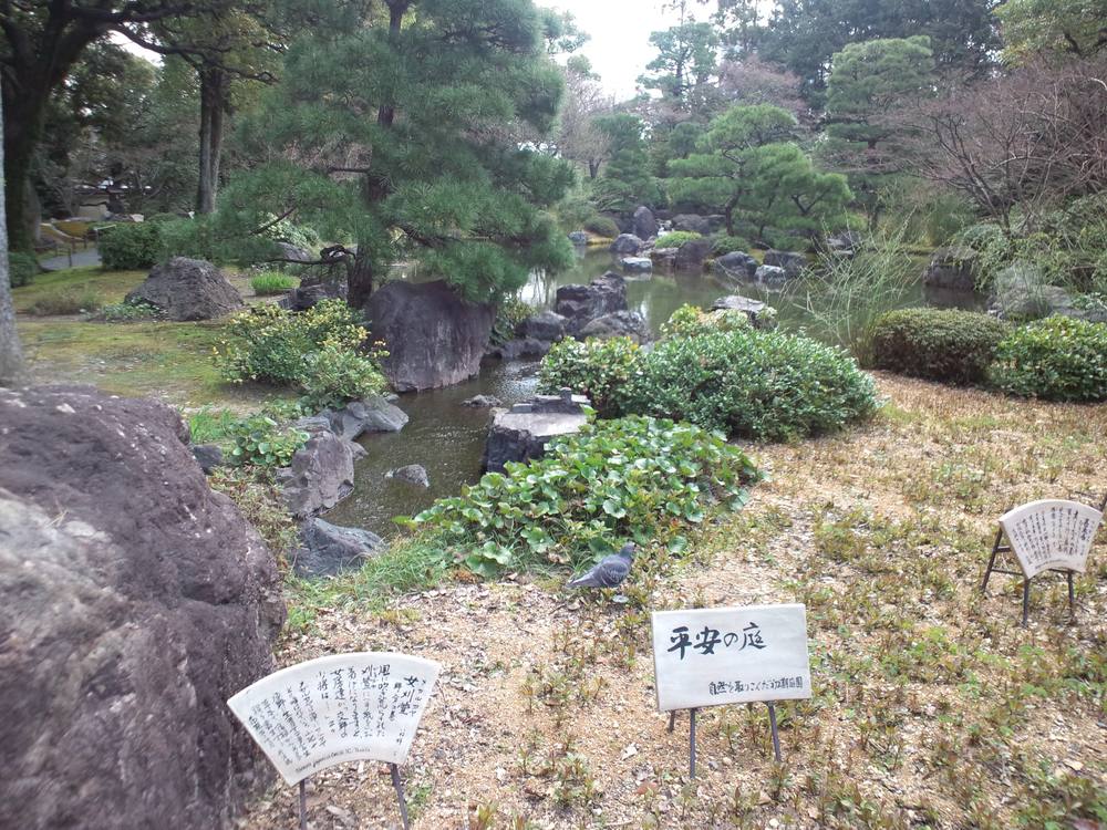 城南宮神苑的平安の庭:池水繞過深色大石與修剪過的松樹,前景地上立著寫有「平安の庭」的白色小牌與兩塊扇形的解說牌,地面還是早春的枯草色