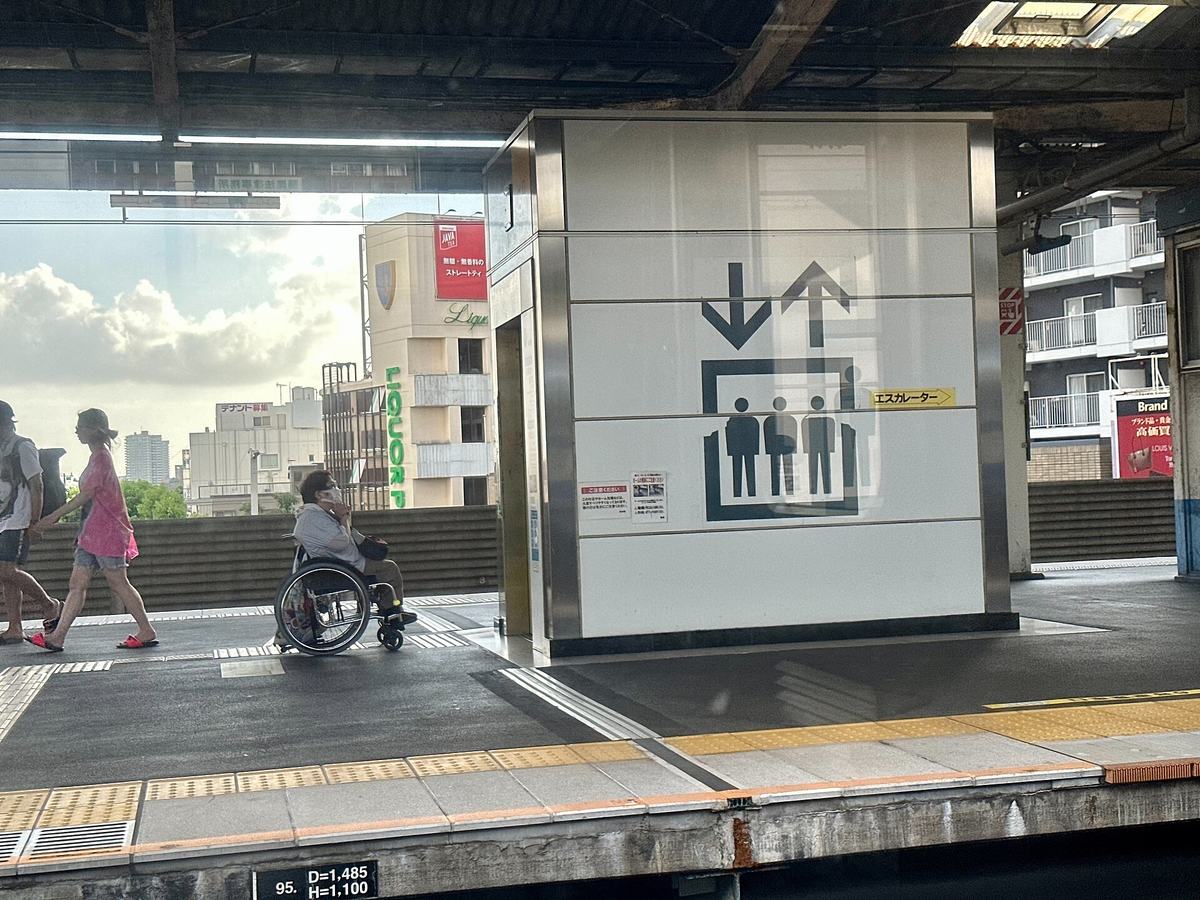 A masked wheelchair user parked alone beside a lift shaft on an elevated Japanese station platform, the shaft wall carrying a large black-and-white lift pictogram and a yellow escalator arrow, yellow tactile paving and the platform edge in the foreground, two pedestrians passing on the left