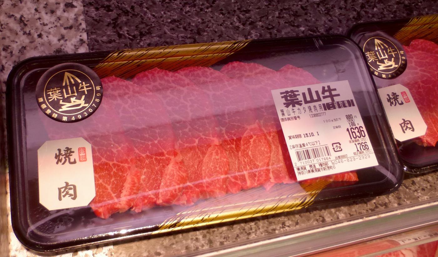 Two cling-wrapped trays of raw beef in a Japanese supermarket chiller: black plastic trays carrying a gold circular Hayama Beef certification seal and a white brush-lettered yakiniku label, with a price sticker on the right listing Hayama beef shoulder for grilling, an individual animal identification number, 880 yen per 100 g, net weight 186 g, 1,636 yen before tax, 1,766 yen including tax, and a best-before date
