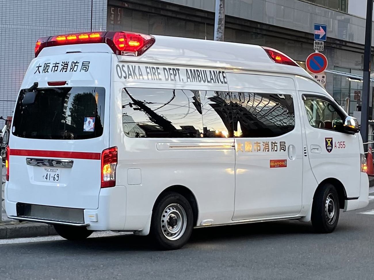 A white Japanese ambulance on a city street with its red roof beacons lit, OSAKA FIRE DEPT. AMBULANCE lettered in black along the upper body, Osaka City Fire Bureau in Japanese above the rear door and on the side, a red stripe along the flank and the unit number A355