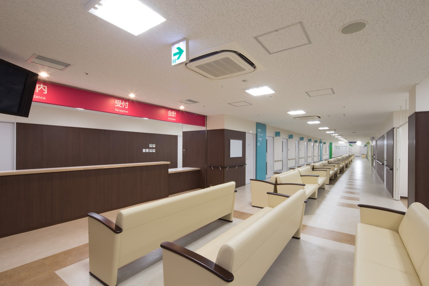 Outpatient hall of a Japanese hospital, a deep red sign band running left to right reading Guidance, Reception and Accounts, each Japanese word carrying one line of English beneath it, a dark wooden counter below and rows of cream leather benches, with a corridor of numbered consulting rooms receding to the right