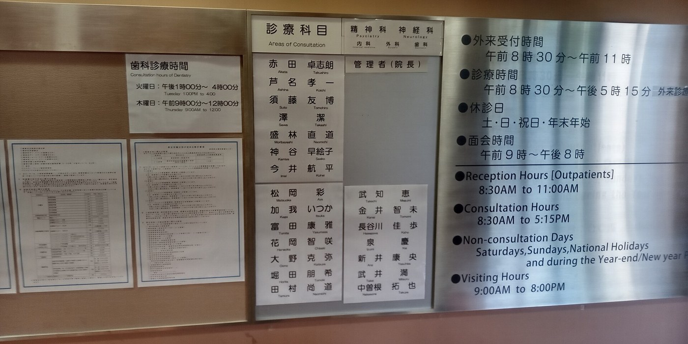 A notice board on the wall of a Japanese hospital: a glazed frame headed Areas of Consultation with a column of doctor names, and a stainless steel plate on the right listing outpatient reception 8:30 to 11:00, consultation hours 8:30 to 17:15, non-consultation days and visiting hours in both Japanese and English