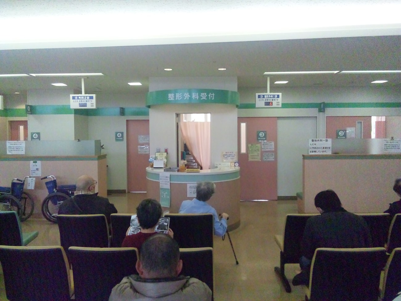 Waiting area of a Japanese hospital: a curved green-topped counter labelled orthopaedic reception in the centre, blue hanging signs on either side for surgery consulting room 2 and orthopaedic consulting room 1 each with a red number display, rows of dark seats with waiting patients, two wheelchairs parked at the left