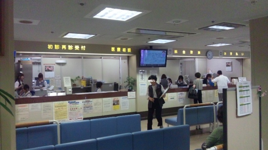 Reception and accounting area of a Japanese hospital, a dark beam overhead carrying yellow Japanese lettering that reads, left to right, first-visit and repeat reception, medical consultation, fee calculation and payment, with a blue queue display and a round clock between them and rows of blue waiting chairs in front