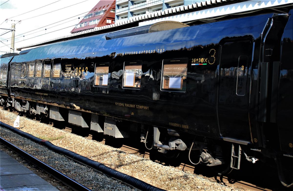 36ぷらす3 一節車廂的側面,黑色車身反射著月台與天空,左半邊是一排緊密的窗、右半邊是三扇較寬的方窗,車門旁漆著金色數字 3,車身寫著 DISCOVER KYUSHU EXPRESS 787 與 KYUSHU RAILWAY COMPANY