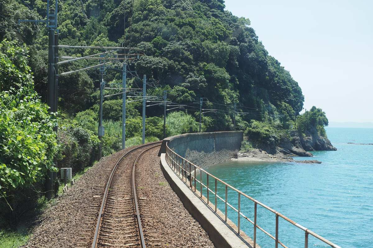 從列車尾端往後看的單線鐵軌,沿著長滿樹的山腳轉彎,右邊緊貼著水泥護岸與欄杆,護岸外是淺藍綠色的海,遠處有岩岸