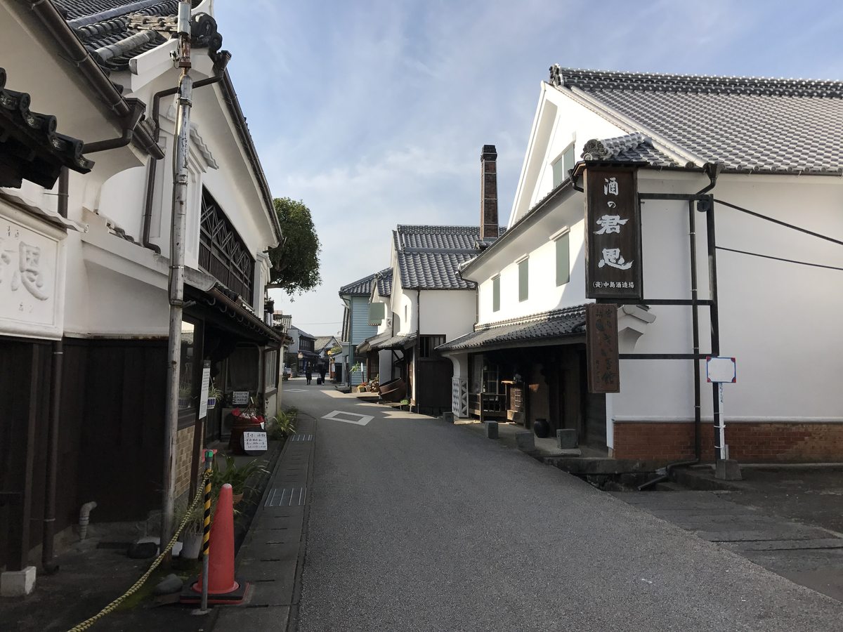 肥前浜宿的酒藏通:一條鋪柏油的窄路兩旁是白牆黑瓦的酒藏,右邊屋簷下掛著直式木招牌,路的盡頭有一根磚造煙囪,遠處有兩三個行人