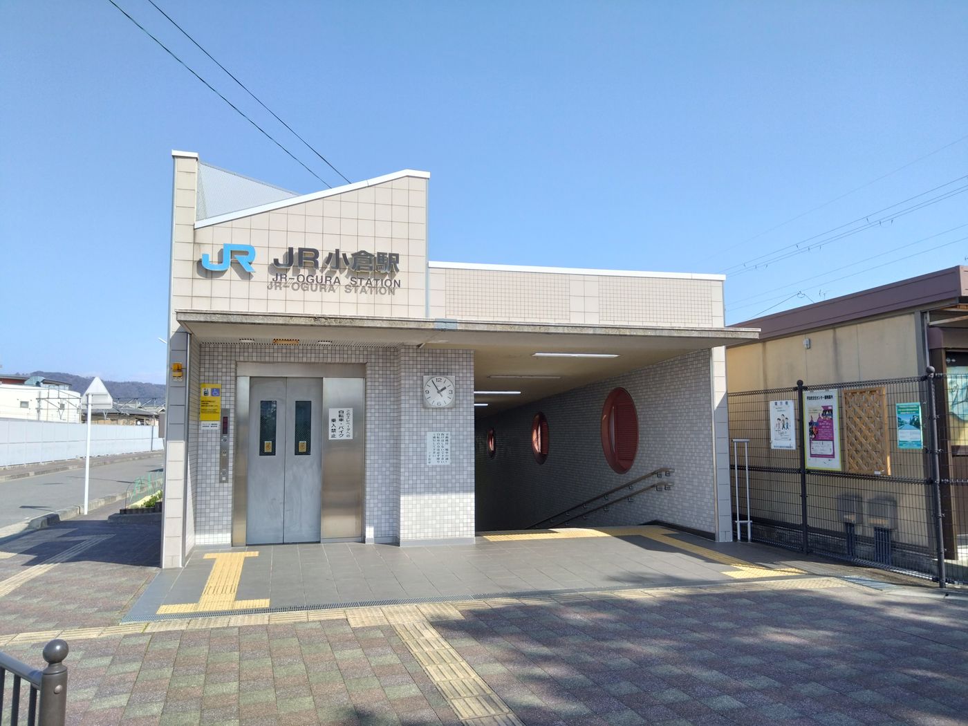 A small station entrance under a clear blue sky: a cream-tiled facade carries the blue JR logo beside the station name in black Japanese characters, with the line JR-OGURA STATION in Roman letters below it; a silver lift door and a round clock face the forecourt, a stairway drops into an underpass on the right, three red circular panels of different sizes are set into the wall, yellow tactile paving crosses grey brick paving, a black fence and a poster board stand to the right, and low houses and a pale ridgeline sit in the distance with nobody in the frame