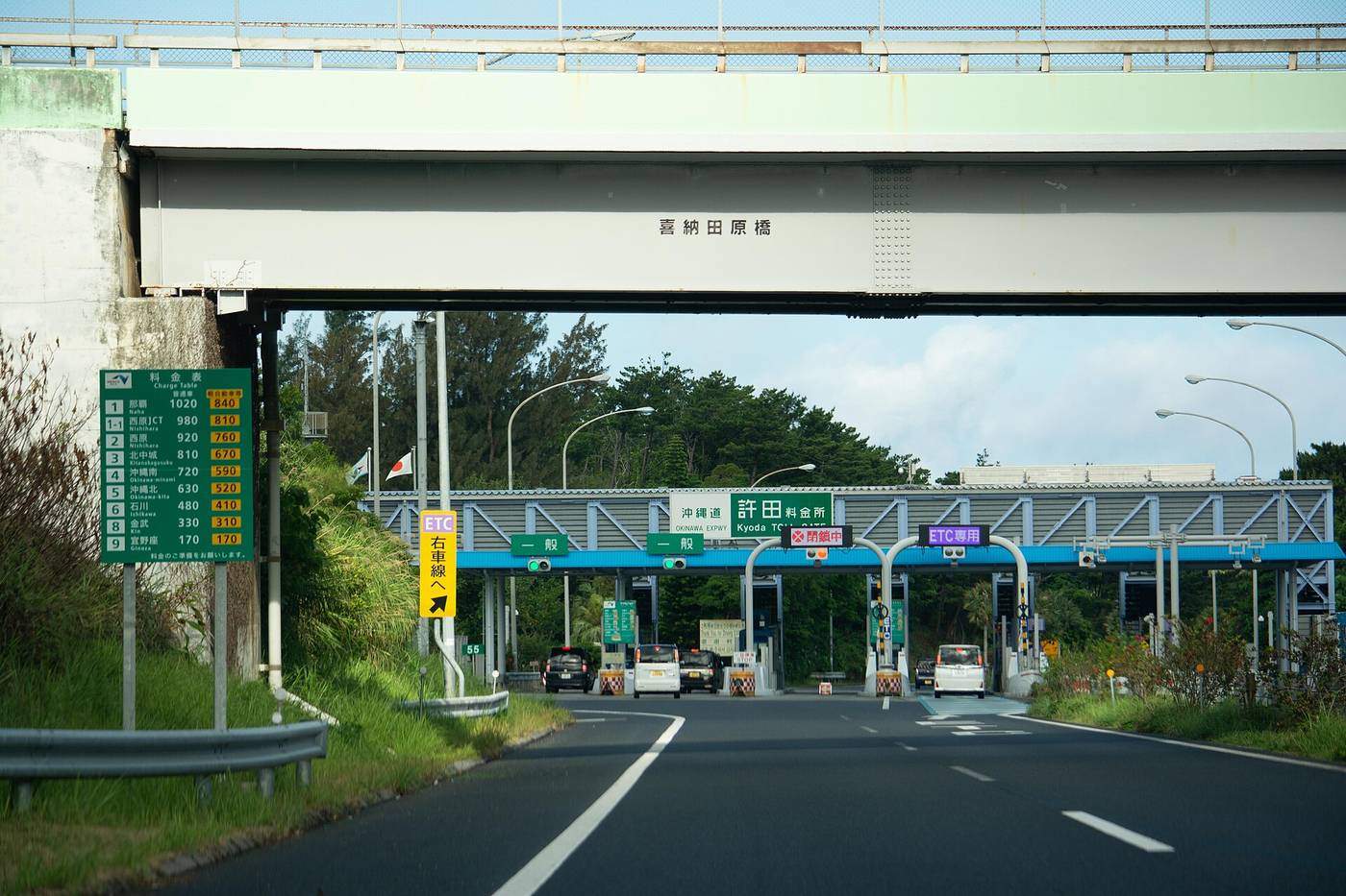 Kyoda toll gate on the Okinawa Expressway seen from a car, a green gantry reading Kyoda TOLL GATE over lanes marked general and ETC only, a green charge table on a post at the left, and an overhead bridge crossing the frame above
