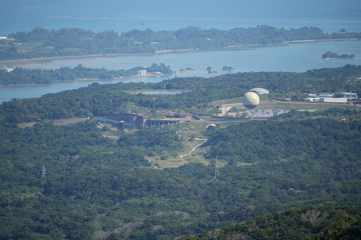 Northern Okinawa seen from Mount Yae: the JUNGLIA site right of centre, ringed by subtropical forest, a cream balloon lettered JUNGLIA beside the brown-roofed buildings and an open lot full of parked cars to its right, with the Haneji inland sea, the far peninsula and open ocean receding behind