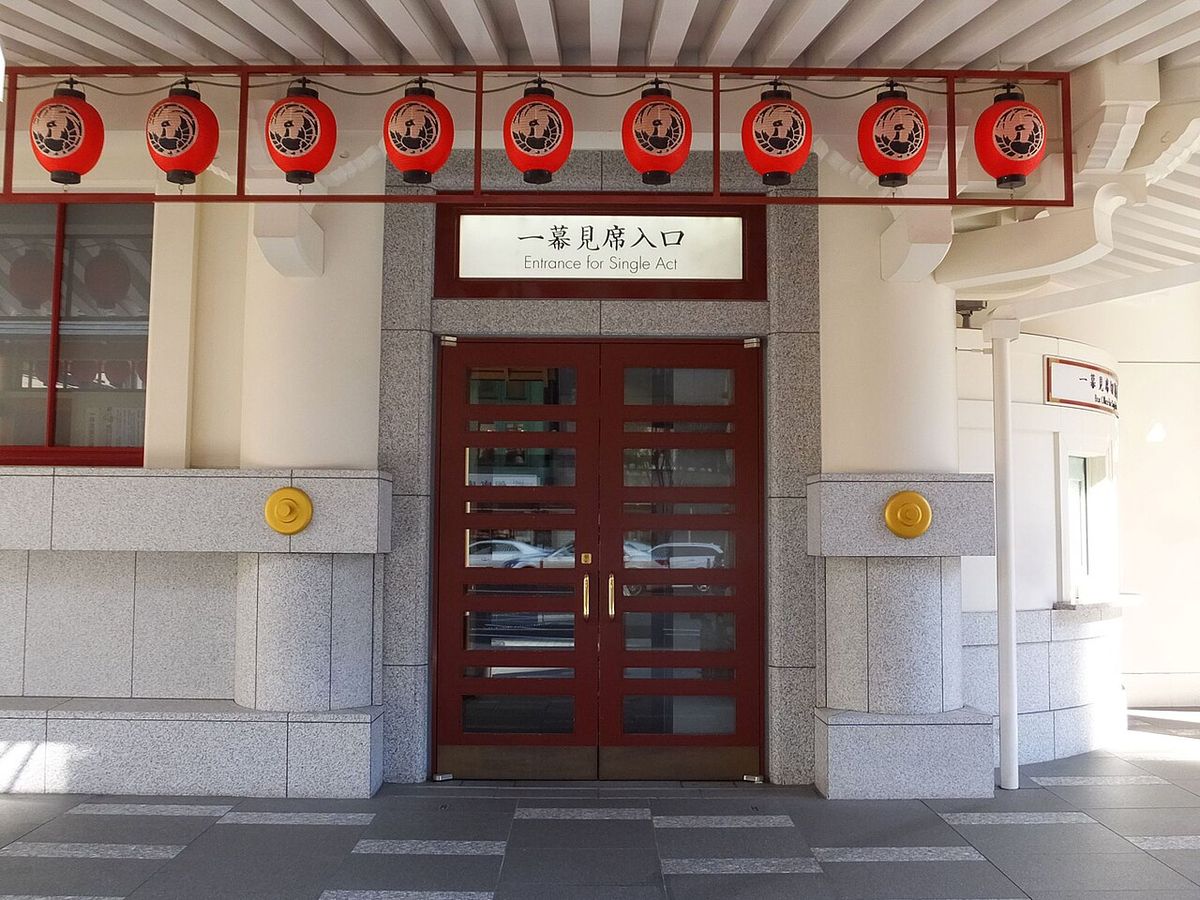 The Kabukiza's single-act entrance: dark red double glass doors under a white sign reading 一幕見席入口 / Entrance for Single Act, with a row of red lanterns bearing the phoenix crest above, and the curved white wall of the single-act box office at the right