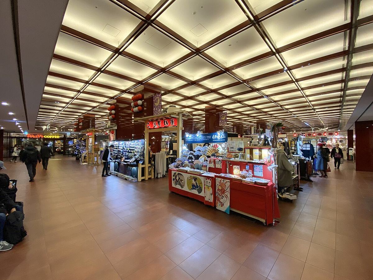 Inside Kobikicho Hiroba on basement level 2 of the Kabukiza: a coffered, backlit ceiling running the length of the hall, a red stall in the centre displaying folding fans and woodblock-print patterned goods, souvenir shops along both sides, a yellow Tully's Coffee sign at the rear left, and red and white paper lanterns hanging from the ceiling