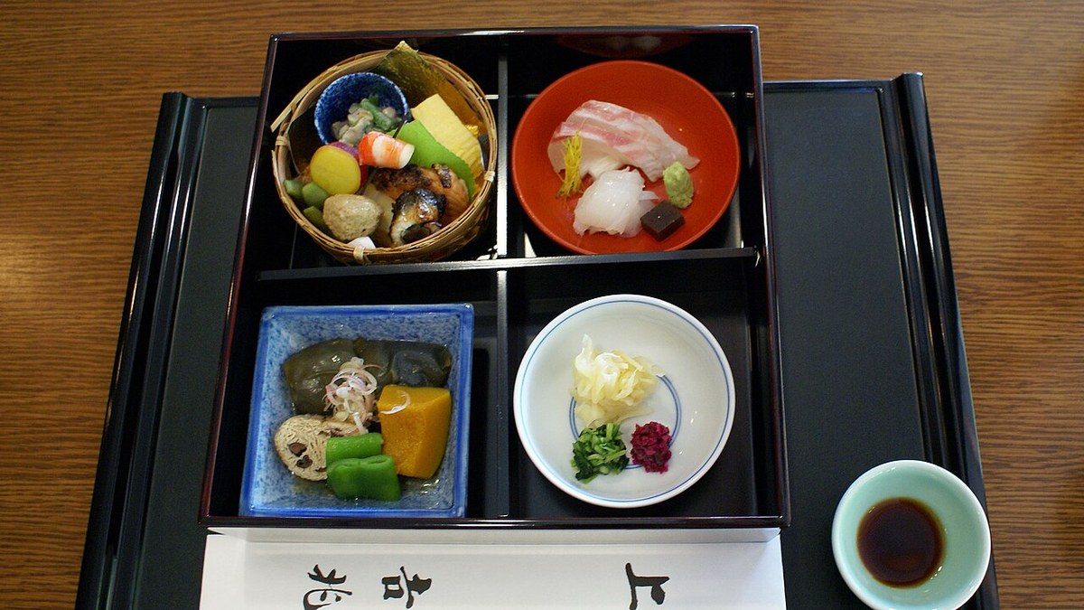 A shokado bento in a black lacquer box divided into four compartments: a woven basket of grilled fish and simmered vegetables, a red lacquer bowl of sashimi with wasabi, a blue square dish of simmered vegetables and a white porcelain dish of pickles, with a small saucer of soy sauce alongside