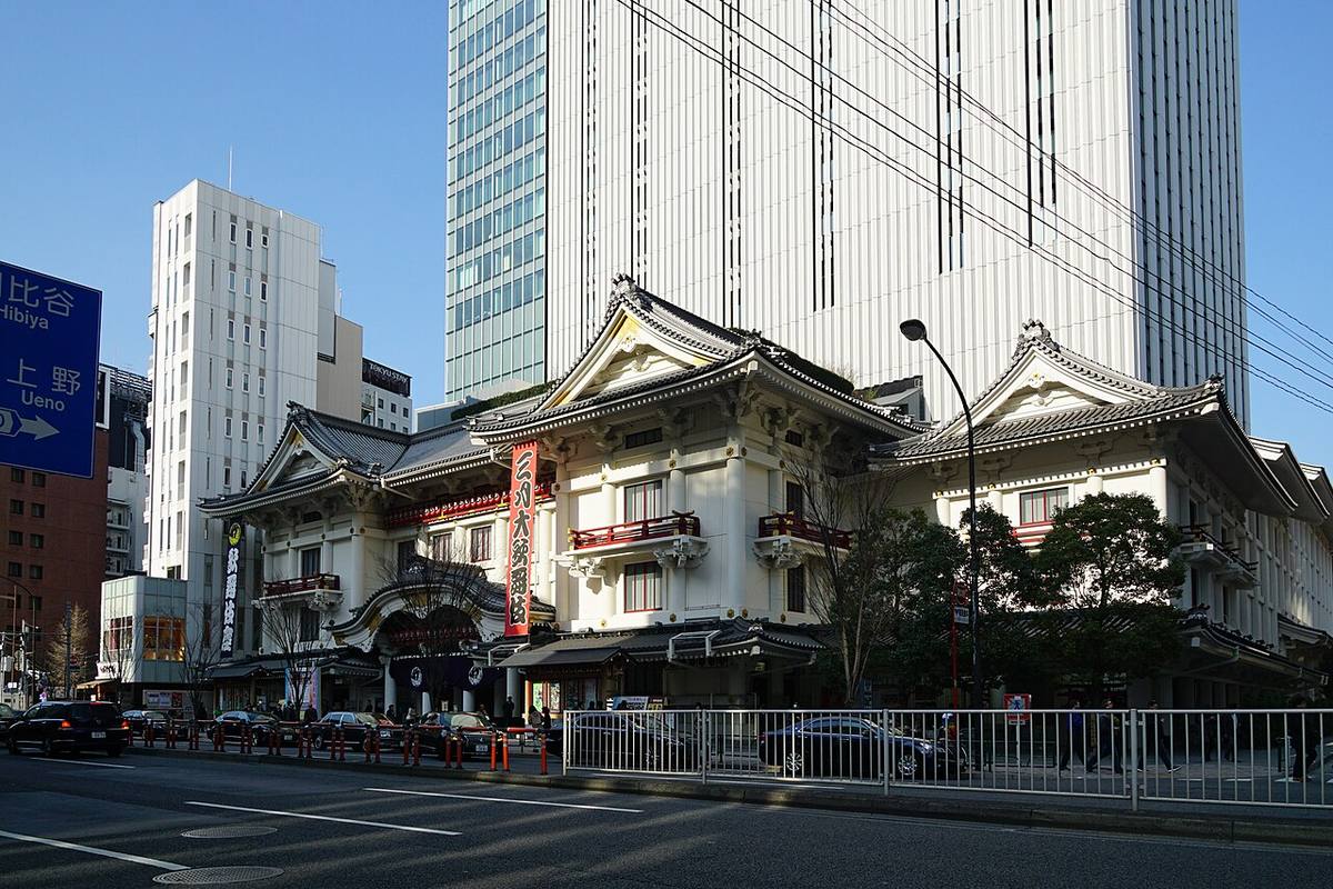 The Kabukiza seen at an angle from across Harumi-dori: three white gabled roofs in a row, a red vertical banner for the current production at the centre, a dark banner at the left end, the grey Kabukiza Tower behind, and a blue road sign for Hibiya and Ueno at the junction