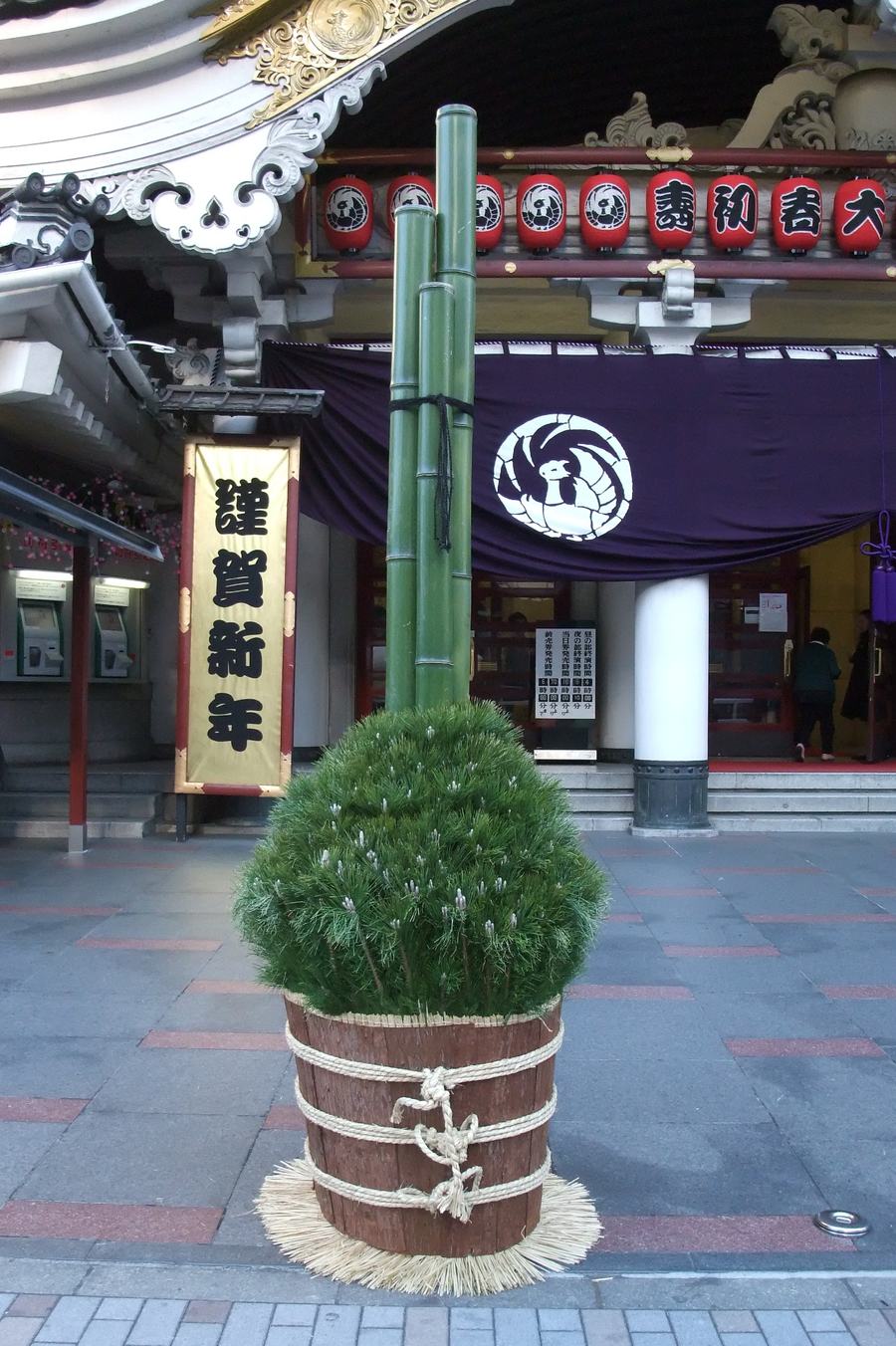 A full-size kadomatsu outside the Kabuki-za theatre: three diagonally cut green bamboo poles set in a rope-bound wooden barrel of pine, with a New Year banner and red lanterns behind