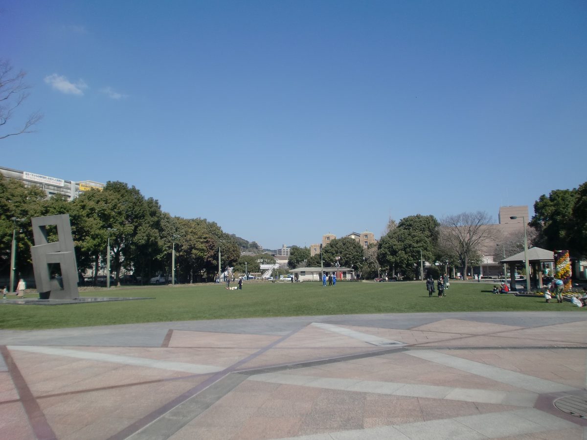鹿兒島市中央公園的大片草坪與石板廣場,左邊有一座灰色方框造型的雕塑,四周是行道樹與低矮建築