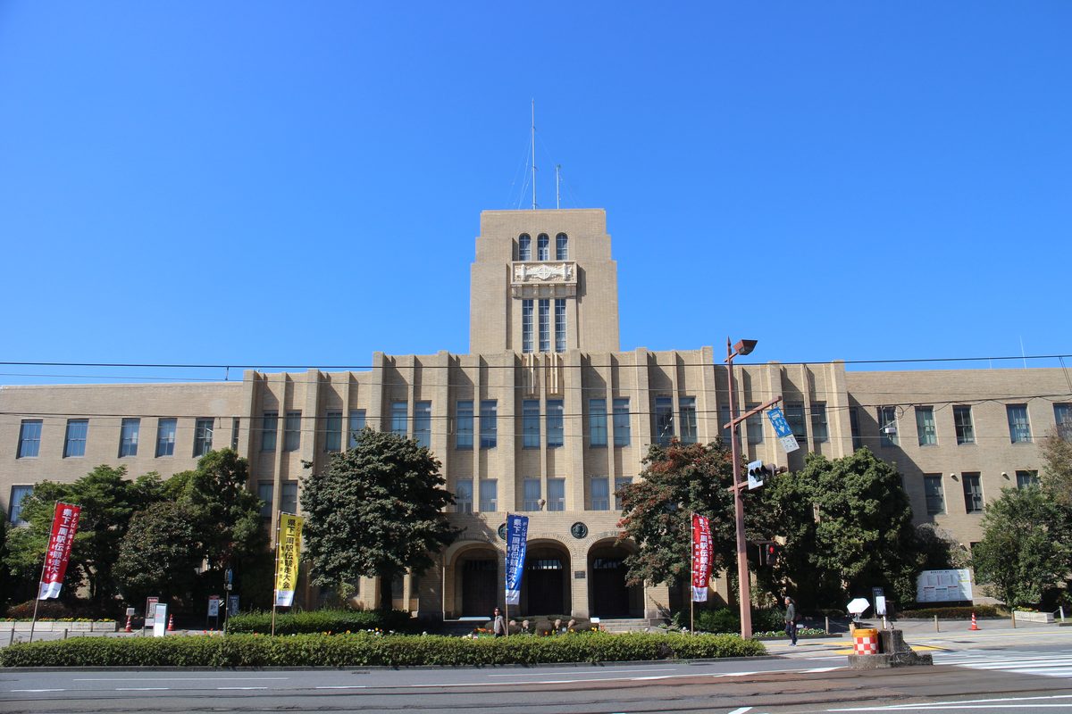 鹿兒島市役所本館正面,米色磚造建築中央有一座高塔與三道拱門,門前插著幾面紅、黃、藍色的直立旗