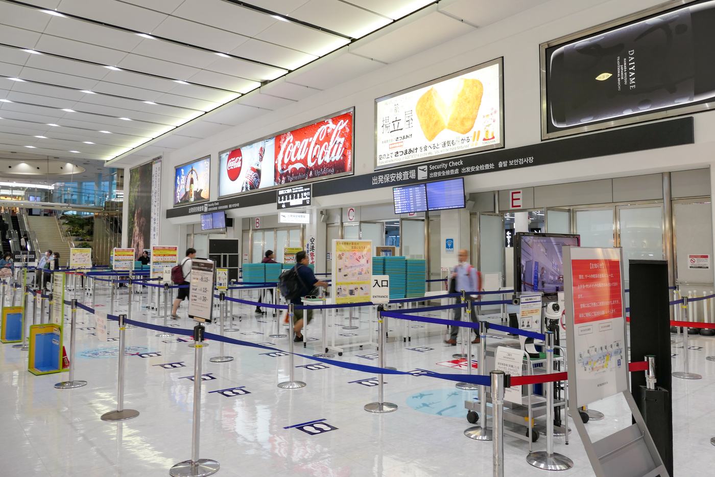 The departure security checkpoint at Kagoshima Airport: a black overhead sign reads "Security Check" in Japanese, English, simplified and traditional Chinese and Korean, lane letters B, C and E are mounted to the right, blue retractable belts mark out the queue on the floor, and stacks of teal trays sit beside the screening tables.