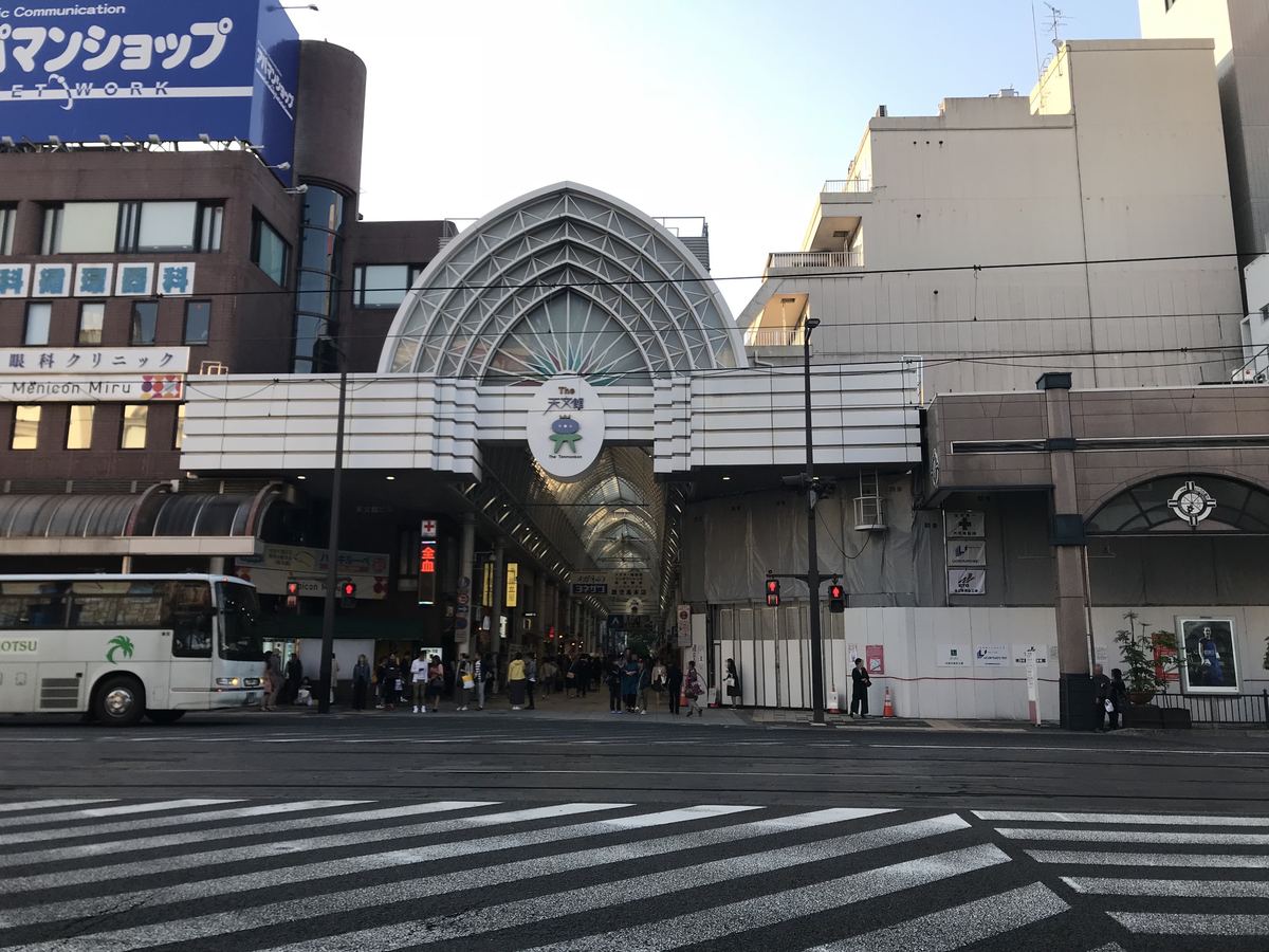 天文館本通り拱廊商店街入口,白色尖拱造型的大門掛著「The 天文館」圓形招牌,門口有行人與一輛白色巴士經過