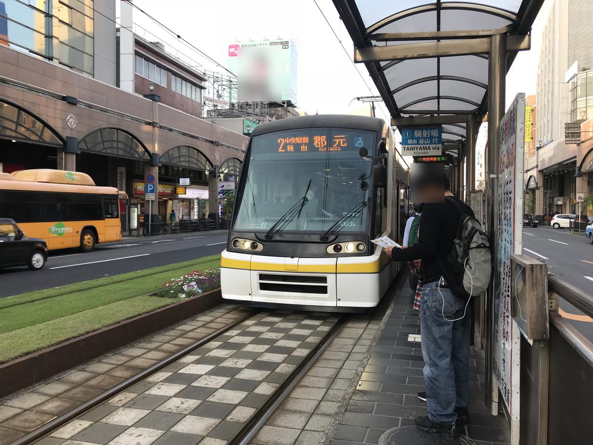 鹿兒島市電天文館通電停,白色與黃色線條的低底盤電車進站,車頭顯示「2 中央駅前経由 郡元」,月台上有乘客等車,臉部已模糊