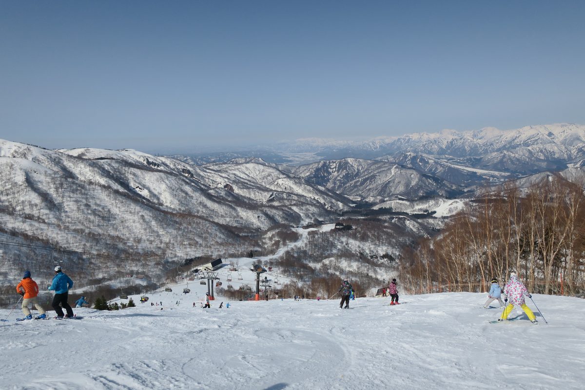 かぐら滑雪場上方的雪道:幾名滑雪者背對鏡頭往下滑,下方山谷有吊椅纜車和山屋,遠方是一層層覆雪的山脈,更遠處是平原