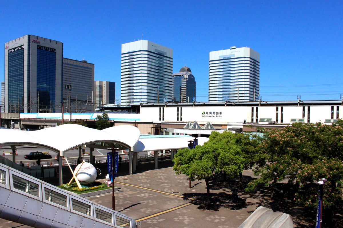 從天橋上看海浜幕張站南口:白色站舍掛著「海浜幕張駅」站名,前面是波浪形白色雨棚與鋪磚廣場,廣場上有一座棒球與球棒的大型紀念物,站後是幾棟高樓