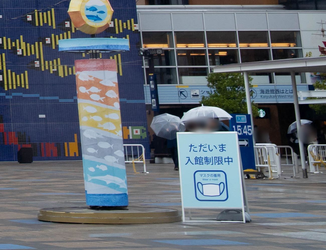 海遊館入口廣場上的淺藍色立牌寫著「ただいま入館制限中」與口罩圖示,後方深藍色看板寫「入館 15:45」,再後面的建築掛著「海遊館西はとば」指標,撐傘的路人臉部已模糊