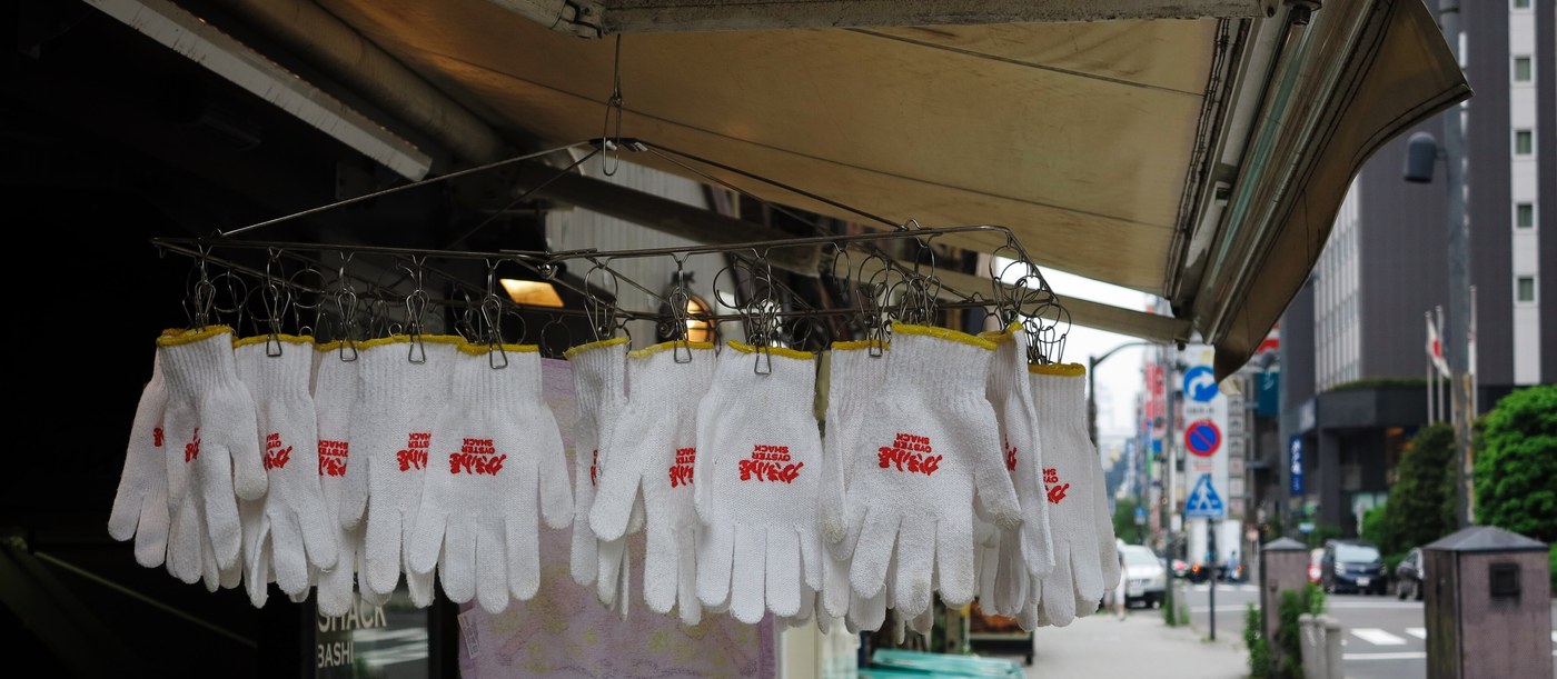 Under a shop awning, a dozen or so white cotton work gloves hang from pegs on a wire drying rack, each printed in red with the words OYSTER SHACK, upside down because the gloves hang inverted; the glass door at lower left shows the words SHACK and BASHI, and a Tokyo pavement with road signs and office blocks runs off to the right