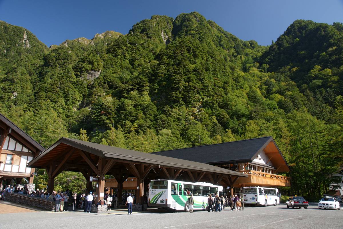 Shuttle buses and queuing passengers under the timber canopy of Kamikochi Bus Terminal, backed by steep forested cliffs