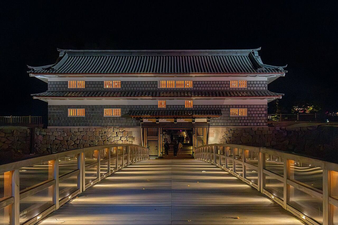 夜間打燈的金澤城鼠多門與鼠多門橋：畫面中央是一座木造拱橋，橋面兩側的扶手下裝著地燈往內照亮橋板，橋的盡頭是黑瓦白牆、下半部貼著黑色海鼠壁的兩層城門，門洞裡透出暖黃色的光