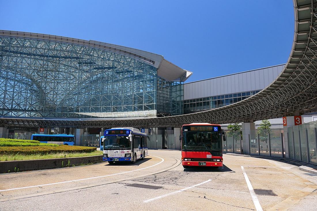 金澤站東口的巴士總站：玻璃帷幕的車站大廳前，弧形屋簷下一排乘車處，柱子上掛著 3、5、6 號的編號牌，兩輛路線巴士正開出來