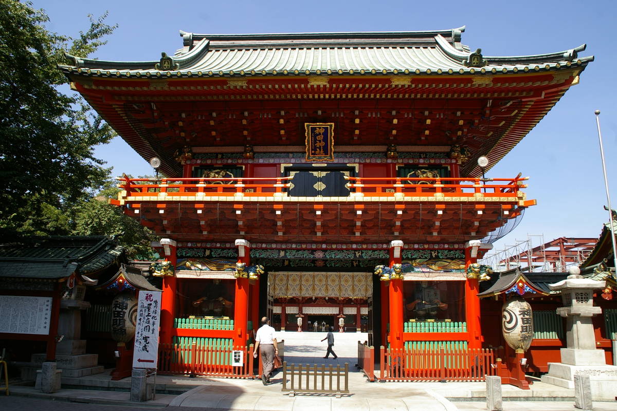 The Zuishinmon gate at Kanda Myojin: a vermilion two-storey gate under a green copper-tiled roof, a dark blue plaque with gold characters beneath the eaves, a large white paper lantern hanging on each side, and the main hall visible through the gateway