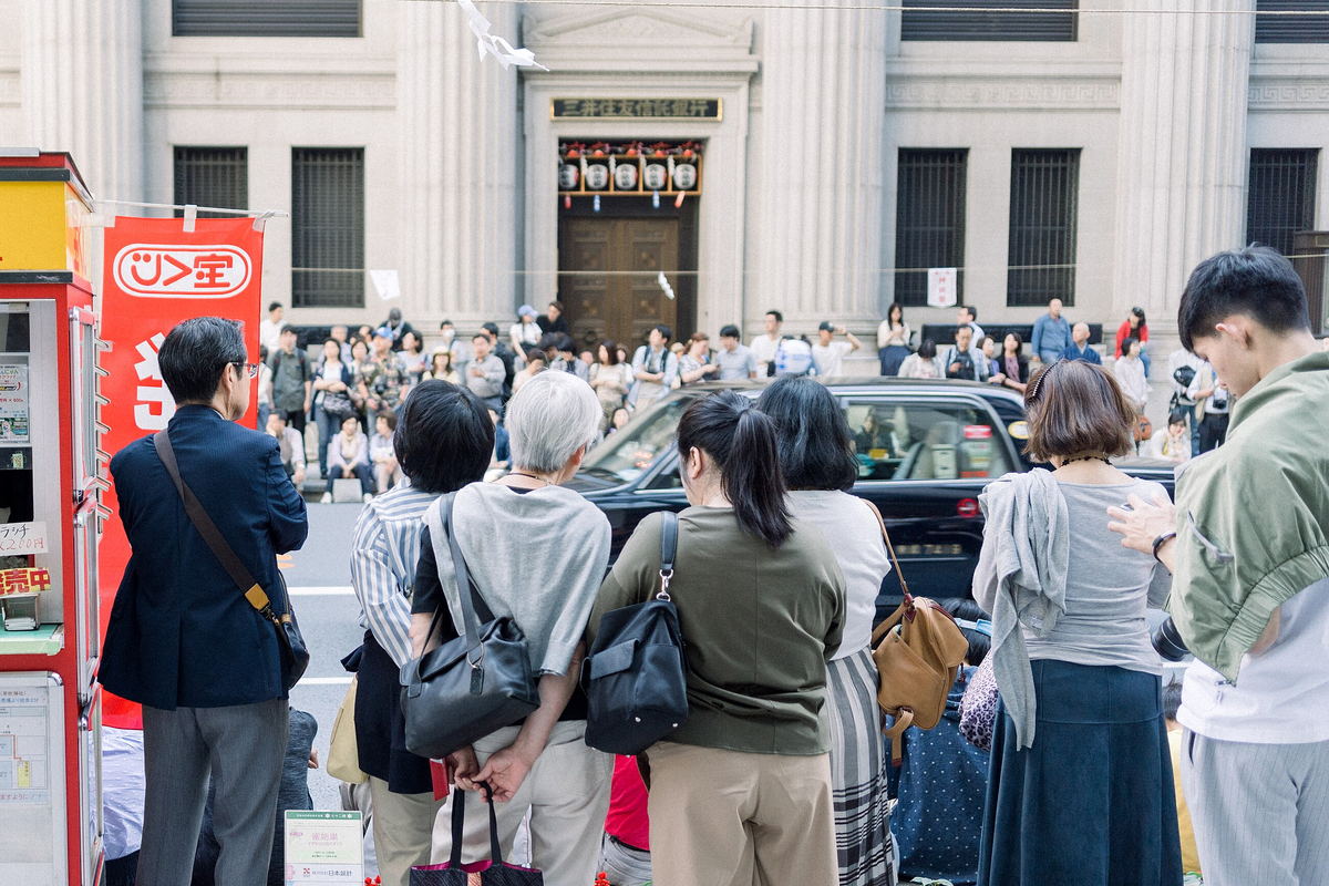 Spectators standing with their backs to the camera on a Nihombashi pavement, waiting; across the road the colonnaded stone frontage of the Mitsui Sumitomo Trust Bank has a row of red-and-white festival lanterns over its doorway and people sitting on its steps, with a black taxi driving past between them