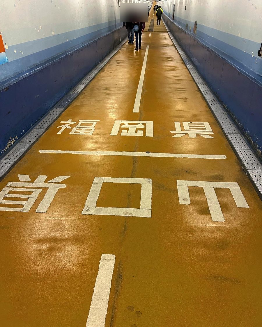 關門隧道人行道的海底通道:黃褐色地面上用白漆寫著「福岡県」與「山口県」,中間一條白線是兩縣的縣界,兩側是藍白色牆面,遠處有幾位行人