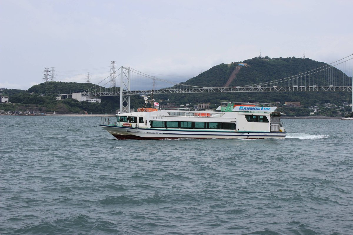 關門汽船的白色雙層客船「かんもん」行駛在關門海峽上,船上掛著 KANMON LINE 招牌,背後是關門橋與綠色山丘,陰天