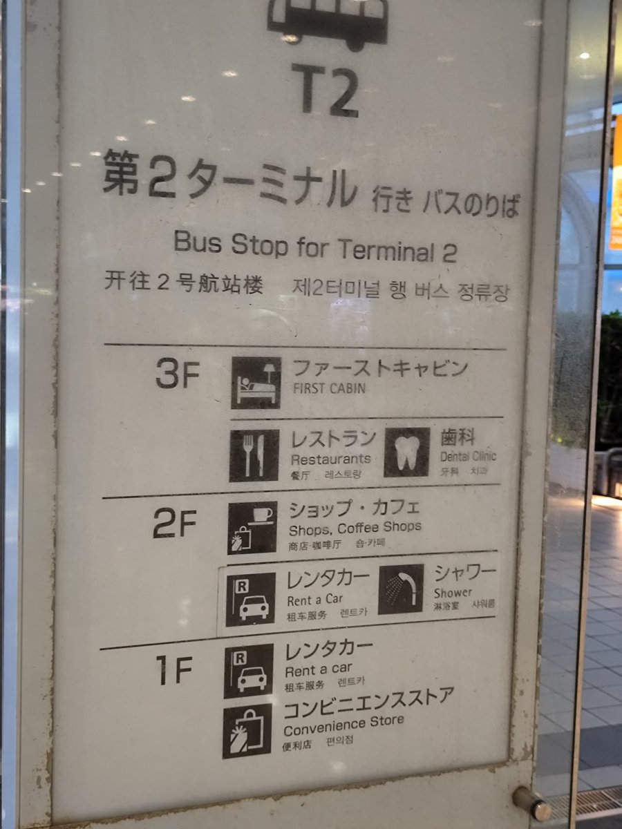 Aeroplaza 的白色樓層看板，最上面是巴士圖示與大字「T2」，下面寫「第2ターミナル行き バスのりば Bus Stop for Terminal 2」與中韓文；再往下列出各樓層：3F 是 First Cabin、餐廳、牙科，2F 是商店咖啡、租車、淋浴，1F 是租車與便利商店