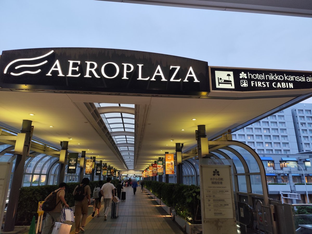 從關西空港站走向 Aeroplaza 的有頂棚連絡天橋，入口上方大招牌寫「AEROPLAZA」，旁邊黑底招牌寫 hotel nikko kansai airport 與 FIRST CABIN；右側立牌是日航飯店的樓層表，背景是飯店大樓，旅客拖著行李往裡走