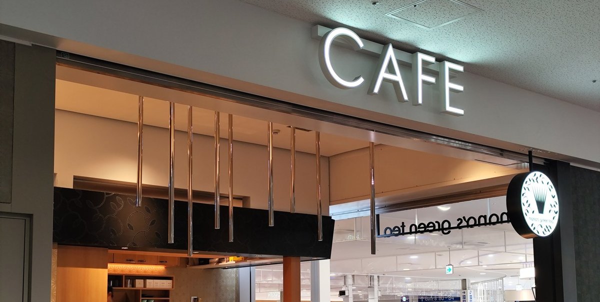 nana's green tea 的店面，入口上方是發光的白色「CAFE」字樣，右側掛著圓形店徽與店名，店內是木質吧台；店外遠處的藍色指標寫著「Gate B 80–84」