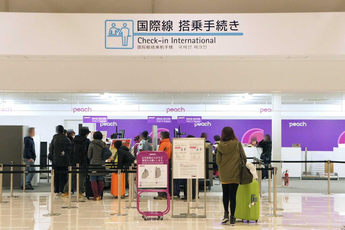 第 2 航廈的國際線報到區，上方白色橫幅寫「国際線 搭乗手続き Check-in International」，下面一整排紫色的樂桃 peach 櫃台，旅客拉著行李在排隊線裡等候，前方立著樂桃的手提行李尺寸確認架